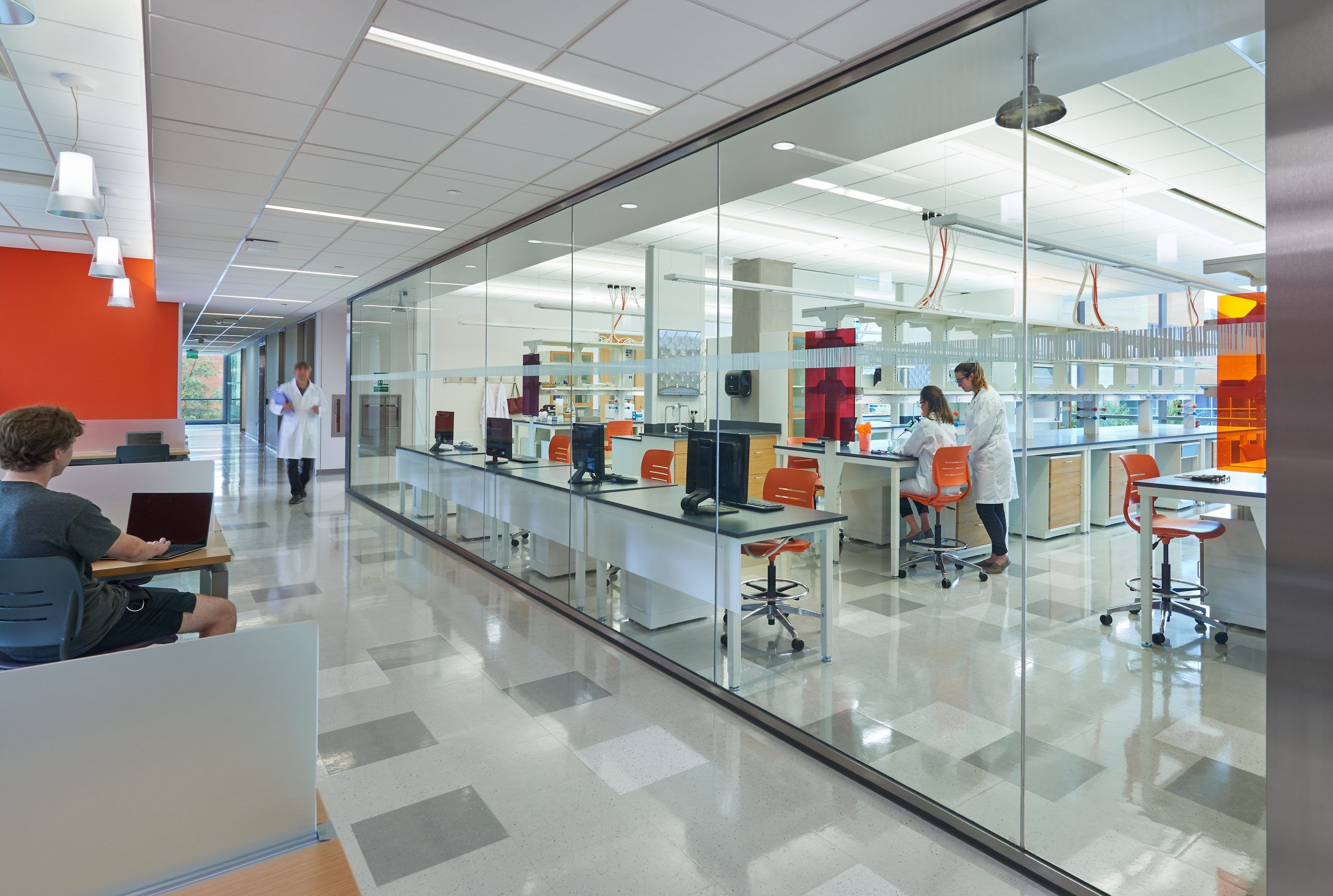 Researchers in a flexible wet lab design with glass walls separating them from students studying in a common area