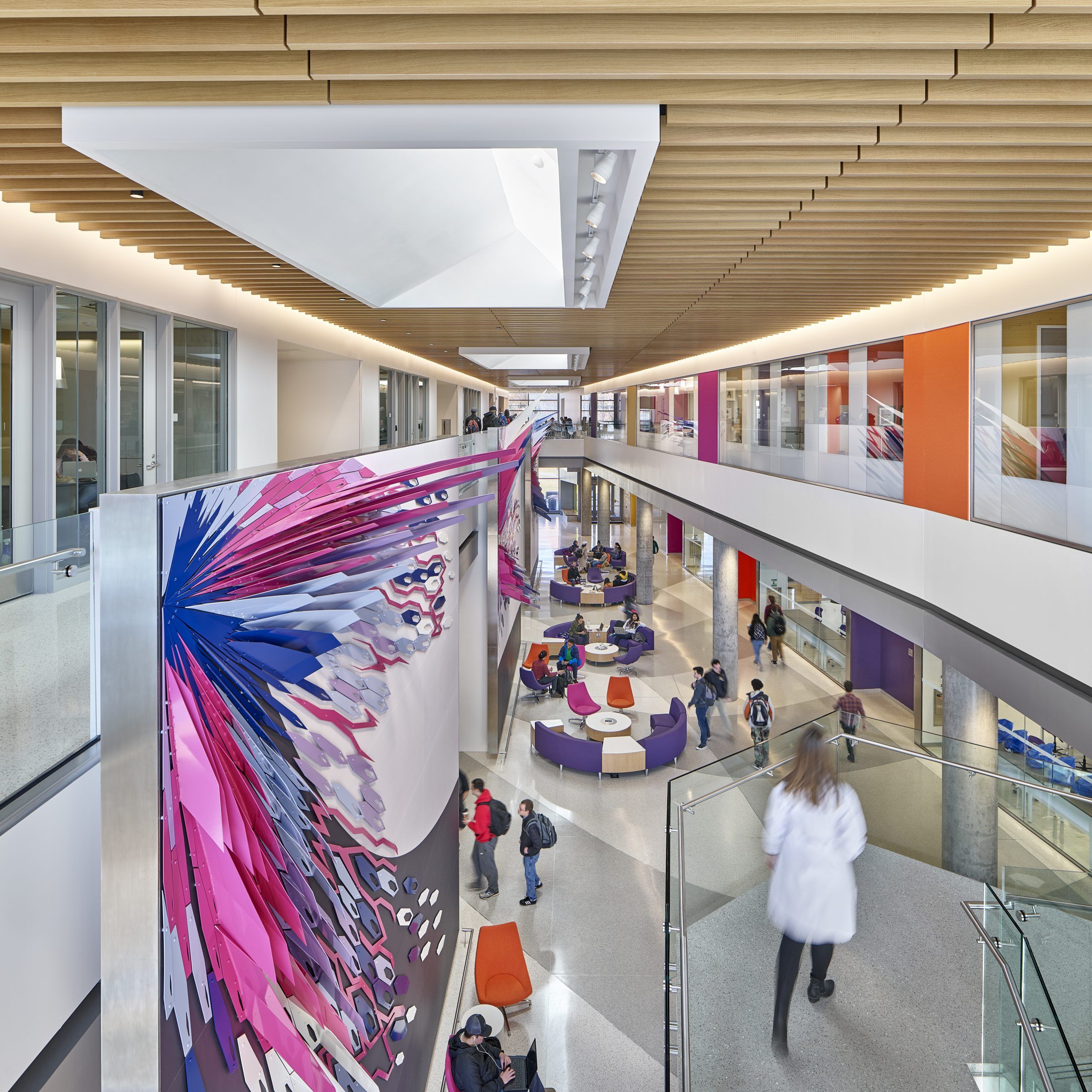 Woman in lab coat overlooking main atrium, with a large purple and pink art installation covering the wall to her left